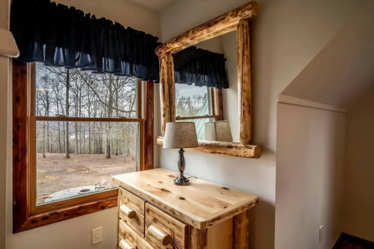 This image features a large wooden mirror, a dresser, and a lamp, with a window that offers a view of the outdoors. The inviting, warm scene hints at the transitional moments between the coziness of the indoors and the beauty just outside the window, especially during the change of seasons.