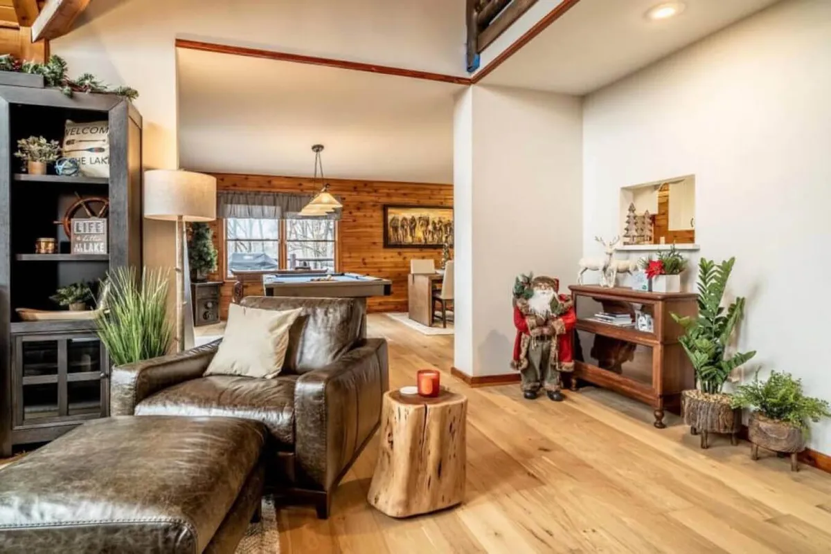 A leather armchair and wooden side table are positioned near a window adorned with curtains and seasonal decorations, including a decorative Santa figure. The scene exudes warmth and coziness, evoking holiday gatherings and intimate winter evenings by the fire.