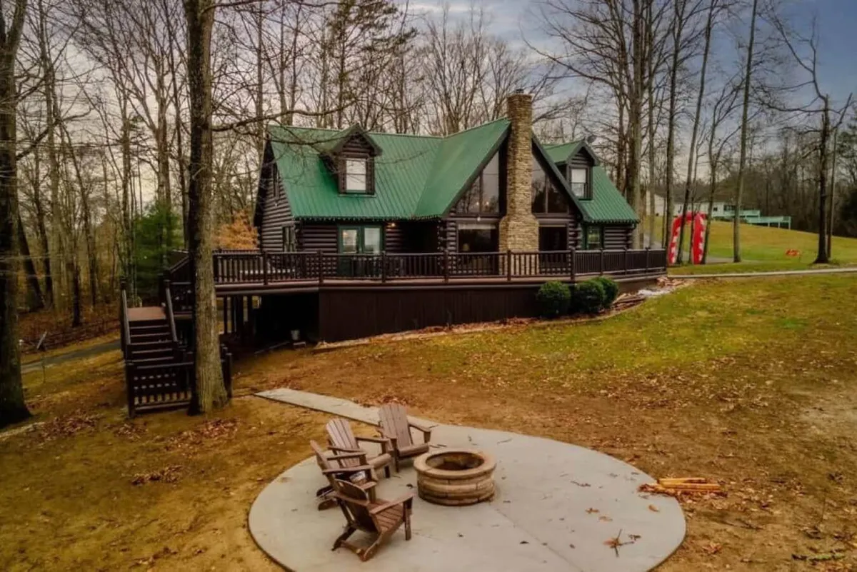 The log cabin’s exterior is shown with a green metal roof and a prominent fire pit encircled by wooden chairs on the lawn. The fire pit setting is ideal for gathering, sharing s’mores, and enjoying evenings under the stars, framed by lush woodland.