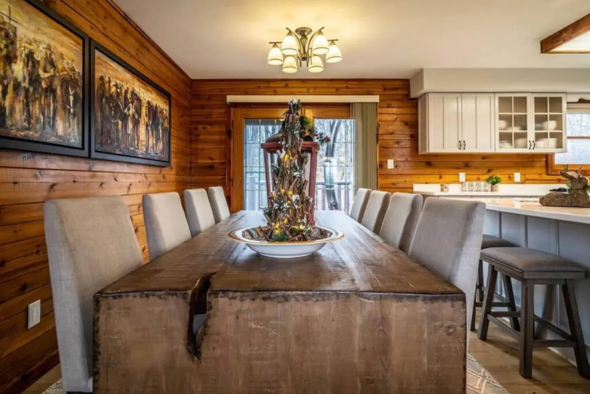 A spacious dining area centered around a large wooden table adorned with a decorative centerpiece. Framed artwork adds charm to the warmly lit room, creating a setting ideal for family dinners, holiday feasts, or casual gatherings.