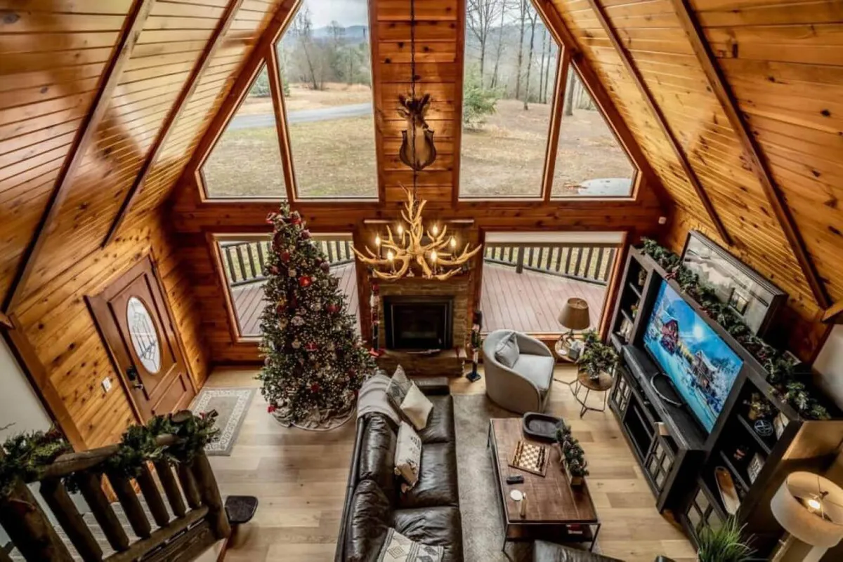 This image captures a spacious living room with large windows, a cozy wood-burning fireplace, an antler chandelier, a leather sofa, and a decorated Christmas tree. The scene is warm and festive, evoking both rustic charm and welcoming luxury—perfect for gatherings, relaxation, and holiday celebrations.