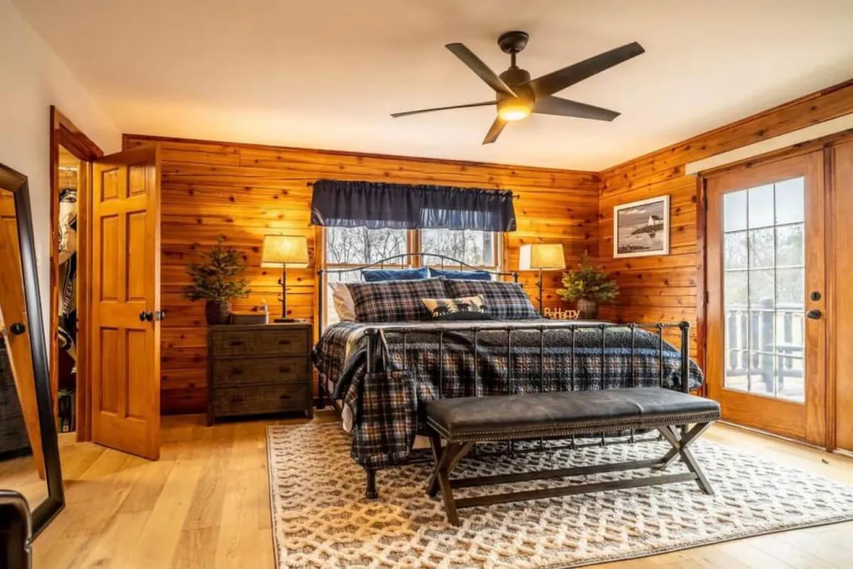 A cozy bedroom featuring a comfortable bed with rustic wooden accents, flanked by nightstands and a bench. The large window allows natural light to stream in, offering tranquil views of the outdoors and creating a restful retreat atmosphere.
