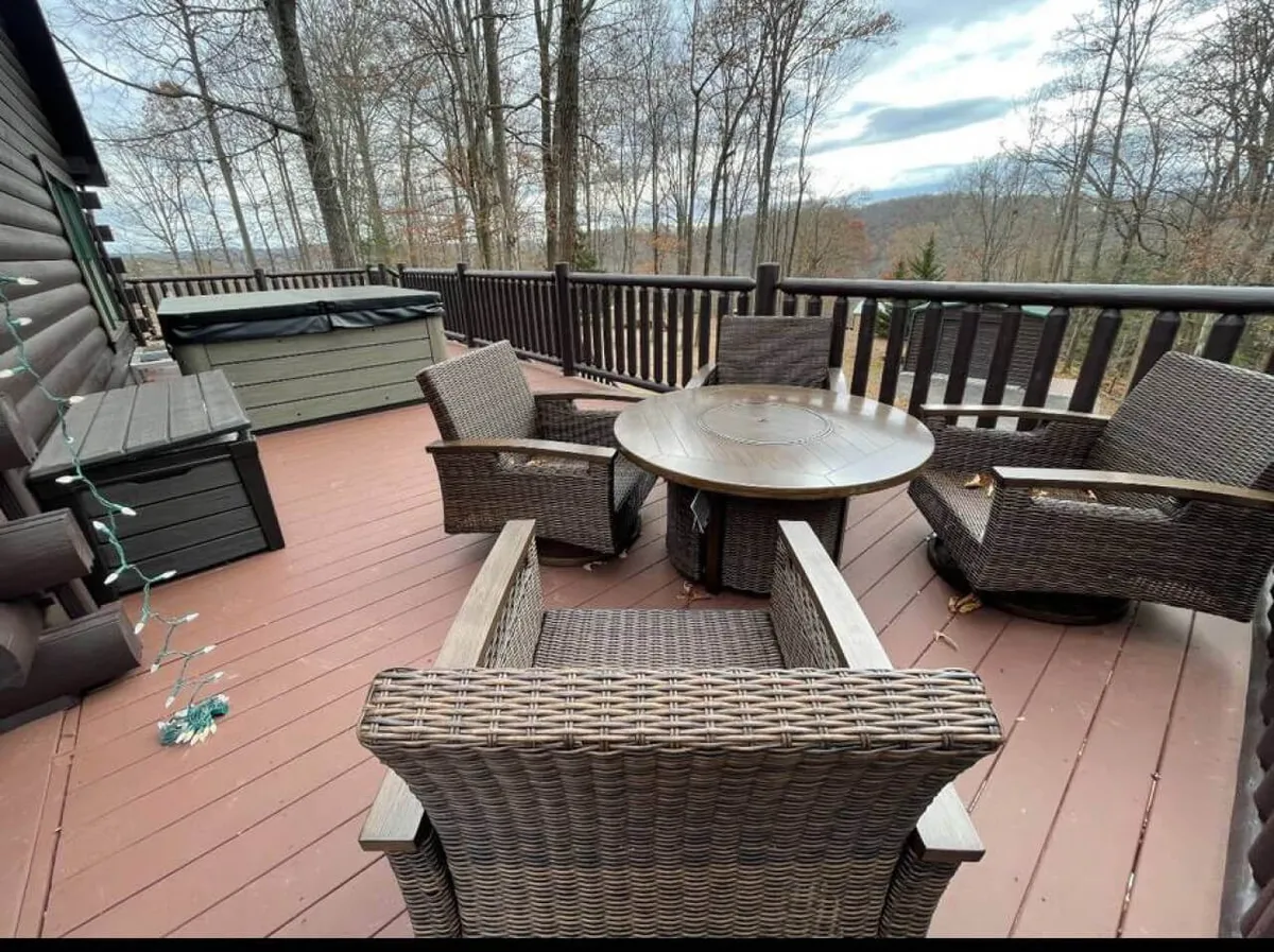 A spacious wooden deck nestled among the trees, featuring wicker chairs, a circular table, and a private hot tub. The surrounding forest creates an intimate, serene atmosphere, perfect for enjoying the outdoors in comfort. Subtle lighting suggests this is a perfect place for evening relaxation or morning coffee.