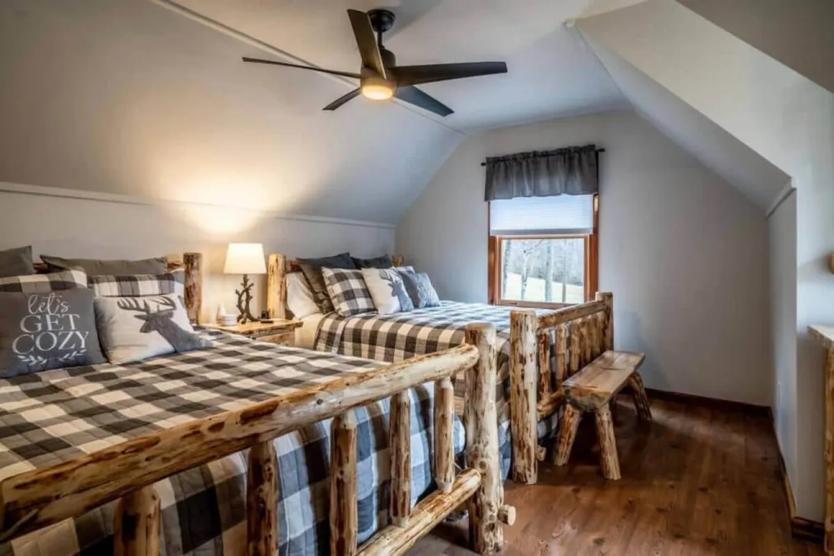 A bedroom with log frame beds and plaid bedding, illuminated by a warm lamp beside a window that frames natural scenery. The room is welcoming, spacious, and clearly designed for guests of all ages.
