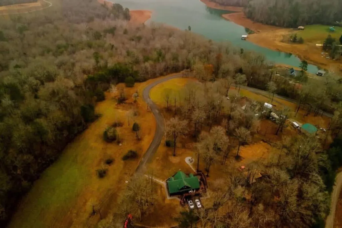 This image captures the green-roofed cabin nestled among tall trees, with a winding road leading toward a tranquil lake visible in the background. The natural surroundings evoke a sense of adventure and relaxation, perfectly tying into the theme of outdoor exploration.