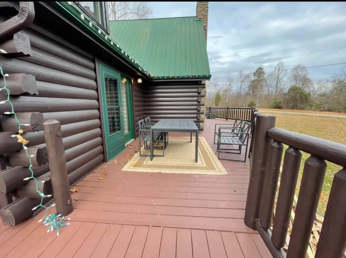 A spacious deck with log cabin detailing offers outdoor seating, a table and chairs, and overlooks the wooded landscape. The setting suggests opportunities for gathering, dining outdoors, or relaxing after a day of lake activities.