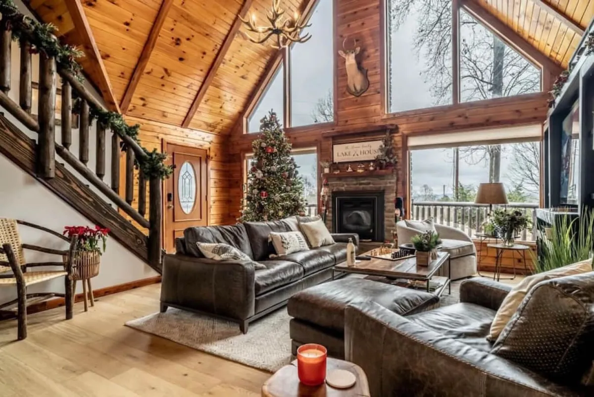 The image showcases the warm, rustic interior of Minnie's Mountain Lake House, featuring a central stone fireplace, plush leather sofas, large windows framing wooded views, a festive Christmas tree, and an inviting wooden staircase. Natural light streams in, creating a cozy space perfect for relaxing, gathering with family, or unwinding with a good book by the fire.