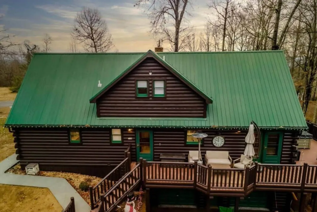 A broad view of the log cabin structure with a green metal roof, a wide wooden deck, and an outdoor seating area. The clock on the wall and the arrangement of chairs invite guests to relax, socialize, and take in the natural surroundings.
