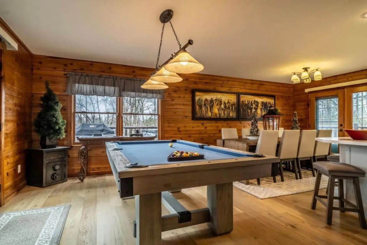 The image features a game room area with a pool table, dining space, large windows letting in natural light, and artwork on the wall. It exudes a relaxed and social atmosphere ideal for game nights and group activities.